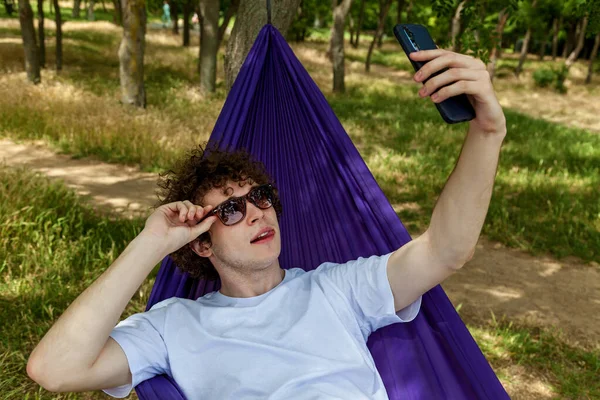 若い男は携帯電話でセルフィーを取り 自然の中で紫色のハンモックでリラックスし 沈黙を楽しんでいます — ストック写真