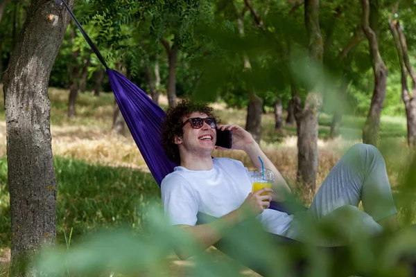 Stock image A young guy is talking on the phone and drinking orange juice while lying in a purple hammock in nature, enjoying the silence.