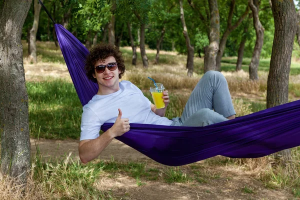 stock image A young guy holds an orange juice with one hand and shows class with the other hand while lying in a purple hammock enjoying nature.