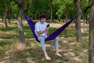 Genç bir adam mor bir hamakta oturur, mor kulaklıkla müzik dinler ve telefonuna bakar... kıvırcık saçlı genç bir adam sıcak bir yaz gününde hamakta dinlenir..