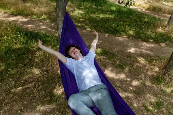 stock image A young guy in a white T-shirt and jeans is lying in a purple hammock and enjoying solitude. A young guy with curly hair is relaxing on a hammock on a hot summer day.