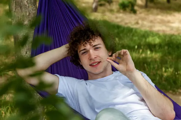 stock image A young guy in a white T-shirt and jeans is lying in a purple hammock and enjoying solitude. A young guy with curly hair is relaxing on a hammock on a hot summer day.