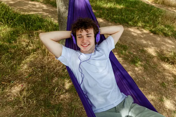 Stock image A young guy is lying in a purple hammock listening to music on purple headphones. A young guy with curly hair is relaxing on a hammock on a hot summer day.