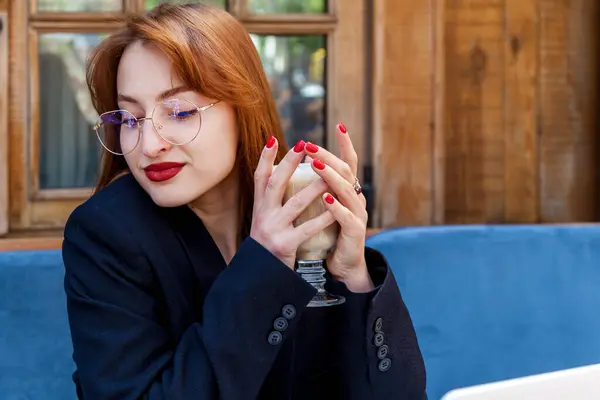 stock image A girl with glasses with red hair in a black jacket sits in a cafe and holds a mug of coffee in her hands. A girl with glasses and red hair with a cup of coffee in her hands