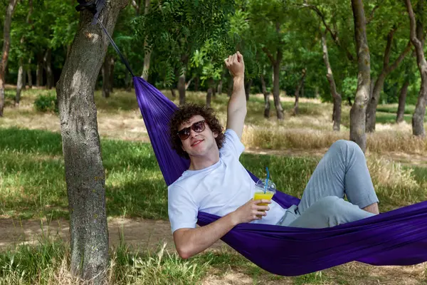 stock image A young guy is lying in a purple hammock drinking orange juice. A young guy with curly hair is relaxing on a hammock on a hot summer day.