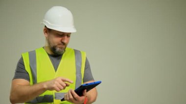 Yüksek görüş açısına sahip güvenlik yeleği ve kasketi olan bir inşaat işçisi tablet bir cihaz kullanıyor. Görüntü, teknolojinin inşaat sektöründeki modern bütünleşmesini, güvenlik protokollerini ve sektördeki dijital araçları vurguluyor. 
