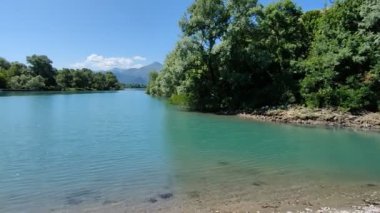 Huzurlu bir turkuaz nehri, berrak mavi bir gökyüzü altında canlı yeşillikler ve uzak dağlarla çevrili verimli bir arazide nazikçe kıvrılır. Sakin su ve doğal güzellik sakin ve sakin bir manzara yaratır..