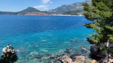 Bu çarpıcı görüntü, arka planda yemyeşil ağaçlar ve görkemli dağlar tarafından çerçevelenmiş kayalık bir kıyı şeridi boyunca canlı mavi suları yakalar. Seyahat, doğa ve açık hava temaları için ideal.