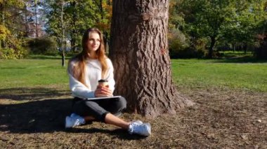 Genç bir kadın güneşli bir günde güzel bir parkta bir ağacın altında otururken dizüstü bilgisayarıyla uzaktan çalışmaktan zevk alır. Yanında bir kahve fincanıyla, odaklanmış ve rahatlamış, üretken kalarak doğayı kucaklıyor. Uzaktan kumanda gibi kavramlar için mükemmel 