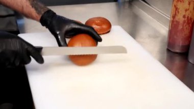 Siyah eldivenli bir şef kesme tahtasında taze bir hamburger ekmeğini dikkatlice keser. Restoran mutfağında hazırlık, işçilik ve kalite kavramı.
