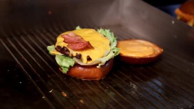 Pişirme işlemi. Izgarada bir şef peynirli ve marullu hamburgerin üzerine çıtır domuz pastırması ekler. Yemek pişirme, yemek blogları veya restoran menüleriyle ilgili video içeriği için mükemmel.