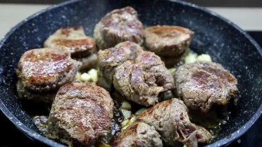 Cızırdayan yağlı ve doğranmış soğanlı tavada pişen yumuşak et madalyonlarının yakın çekimi, altın-kahverengi bir kabuk ve zengin bir doku sergiliyor.