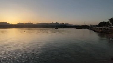 Altın saat manzaralı sakin su, yumuşak sabah ışığını yansıtan, dağ siluetleri ve sahil manzaralı tatil köyü.