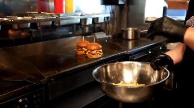 Soslu iki sulu çizburger sıcak ızgarada otururken, siyah eldivenli bir aşçı yakınlardaki bir metal karıştırma kabında patates kızartması pişirir.