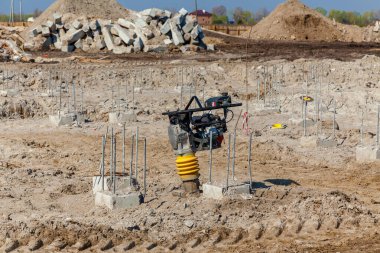 Açığa çıkmış beton zemini, dikey çelik takviye çubuğu ve ön planda toprak sıkıştırıcısı olan inşaat alanı toprak yığınları ve kırık beton bloklarla çevrili.