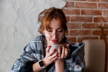 Kırmızı manikürlü, elinde fincanla, sıcak, kırsal bir kafede ekose battaniye ve gözlük takan gülümseyen bir kadın sahnesi.