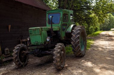Paslı, yeşil traktör. Büyük çamurlu lastikleri var. Kırsal bir yerde, ahşap bir ahırın yanında duruyor.
