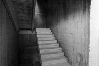 Modern bir inşaat projesinin loş, tamamlanmamış iç kısmında çiğ beton merdiven, açık yüzeyler ve yapısal elementler sergileniyor.