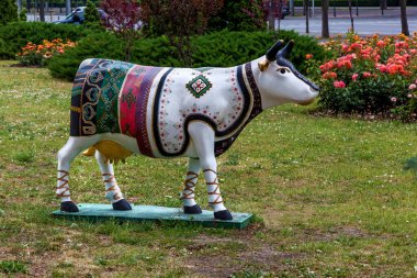 Canlı etnik tasarımlarla boyanmış sanatsal inek heykeli. Şehir parkında çiçek açan çimenlerin üzerinde duruyor.