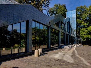 Çağdaş yapı, geniş yansıtıcı pencereler ve parlak granit cepheli, yemyeşil ve açık mavi gökyüzü ile çevrili, parlak ve huzurlu bir açık hava manzarası yaratıyor.