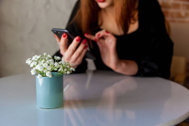 Küçük mavi bir vazoda, parlak bir masanın üzerinde, siyah elbiseli bir kadının akıllı telefonuyla etkileşim halindeki bulanık arkaplanı olan narin beyaz çiçeklerin yakın çekimi.