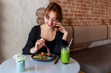 Gözlüklü neşeli genç kadın, duvarları tuğlalı, samimi, modern bir kafede sohbet ederken taze bir yemek ve yeşil bir smoothie içiyor.