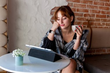 Ekose şallı düşünceli genç bir kadın yuvarlak bir masada oturuyor, elinde bir fincan tutuyor ve tuğla ve alçı duvarlı şık bir kahve dükkanında dijital tablete bakıyor.