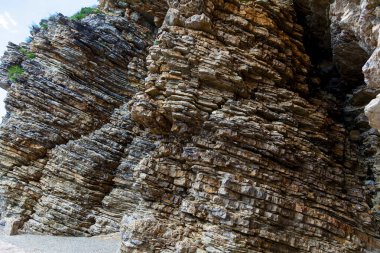 Keskin yatay katmanlar ve engebeli dokuları gösteren katmanlı bir kaya oluşumunun yakın görüntüsü, bir kıyı veya kanyon ortamında doğal jeolojik desenleri ve toprak tonlarını vurguluyor.