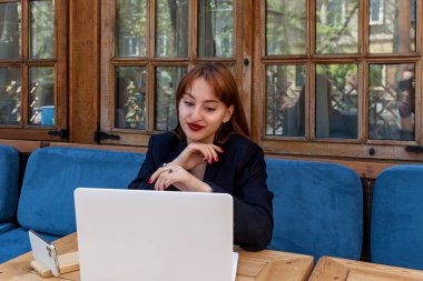 Siyah ceketli genç bir kadın ahşap bir masada beyaz bir dizüstü bilgisayarla oturuyor, uzaktan gülümsüyor mavi yastıklı koltukları ve ahşap çerçeveli pencereleri olan bir kafeteryada çalışıyor.