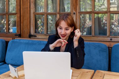Koyu renk ceketli neşeli genç bir kadın dizüstü bilgisayardaki bir video aramasında gülümserken mavi koltuklu, ahşap desenli, rahat bir kafede oturuyor.