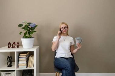 Modern bir iç mekanda oturan, elinde nakit ve telefon yelpazesi olan, düşünceli ya da şüpheci görünen sarışın bir kadın.