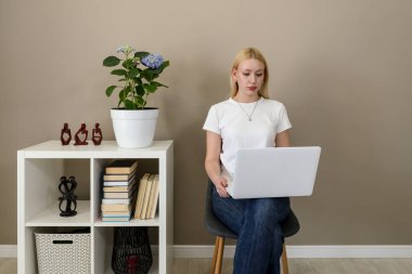 Kucağında dizüstü bilgisayarla bir sandalyede oturan beyaz tişörtlü ve kot pantolonlu sarışın bir kadın kitaplarla, dekorlarla dolu bir rafın yanında işe odaklanmış.