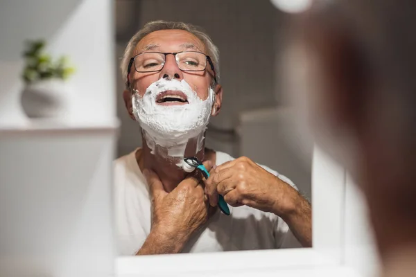 stock image Senior man is shaving his beard with razor and shaving foam in bathroom.