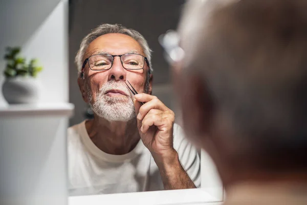 stock image Senior man is picking noise hair with tweezers in bathroom.