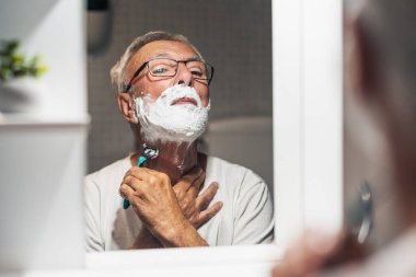 Kıdemli adam tıraş olurken tıraş köpüğü ve tıraş bıçağıyla sakalını tıraş ediyor..