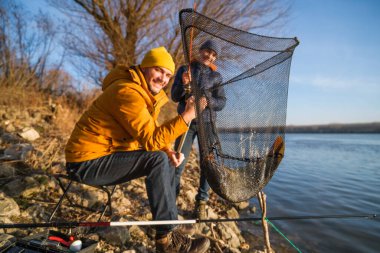 Father and son are fishing on sunny winter day. They caught a fish and are holding it in a landing net.