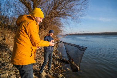 Father and son are fishing on sunny winter day. They caught a fish and are holding it in a landing net.