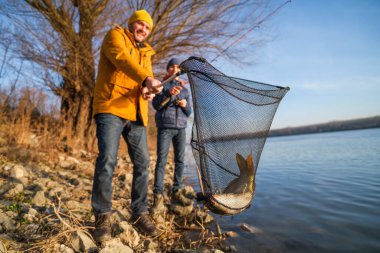 Father and son are fishing on sunny winter day. They caught a fish and are holding it in a landing net.