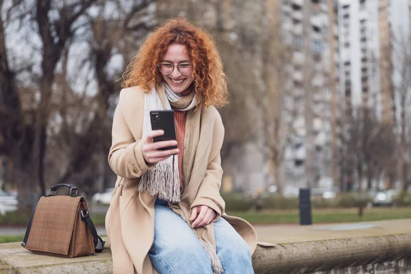 Natural portrait of a caucasian ginger woman with freckles and curly hair. She is smiling and using smartphone.