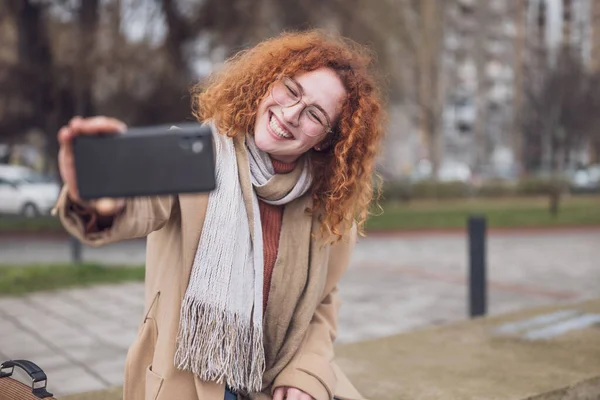 Natural portrait of a caucasian ginger woman with freckles and curly hair. She is smiling and taking selfie.