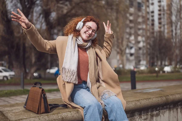 Natural portrait of cheerful caucasian ginger woman with freckles and curly hair. She is listening music and enjoying sun.