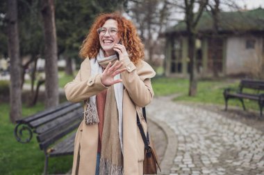 Gülümseyen, kızıl saçlı, çilli, kıvırcık saçlı bir kadının doğal portresi. Telefonla konuşuyor..