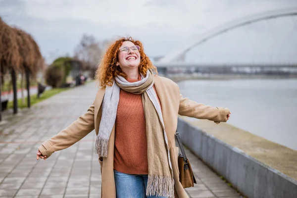 Happy ginger woman with freckles and curly hair is walking on the quay after work. Smiling woman outdoor.