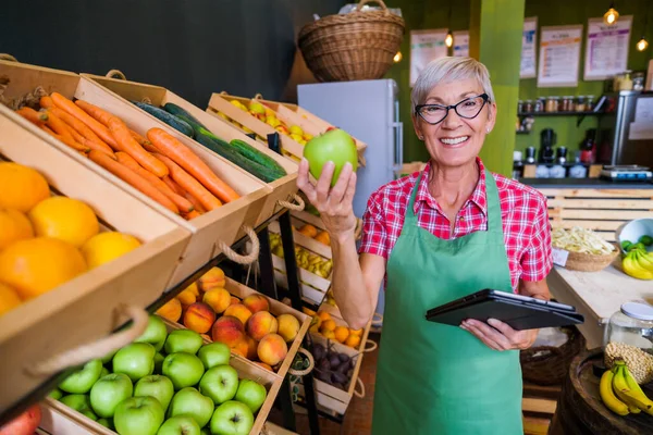 Mature woman is working at fruits and vegetables shop She is examining apples.