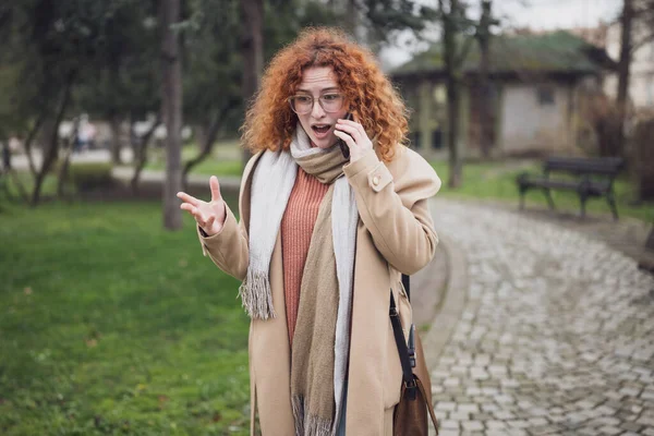 Natural portrait of caucasian ginger woman with freckles and curly hair. She is talking on phone.