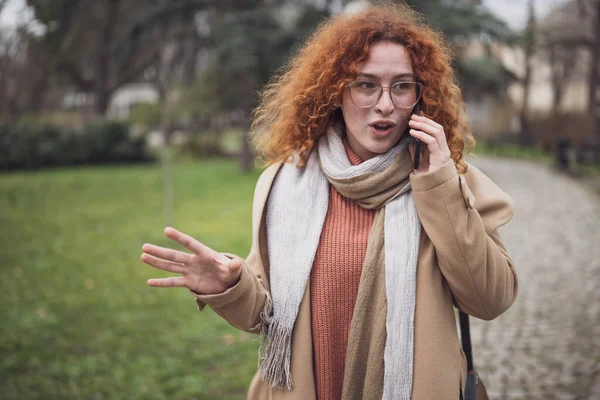 Natural portrait of smiling caucasian ginger woman with freckles and curly hair. She is talking on phone.