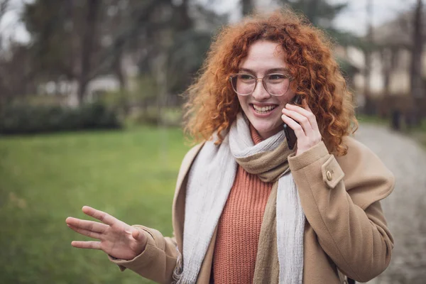 Natural portrait of smiling caucasian ginger woman with freckles and curly hair. She is talking on phone.