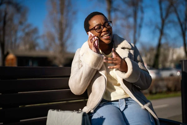 Outdoor portrait of black woman on sunny day. She is sitting on bench in the street and talking on phone.