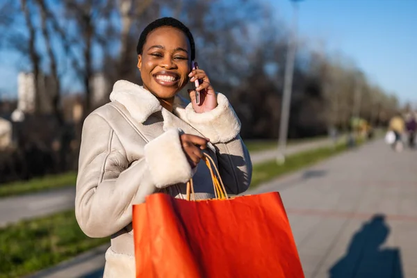 Outdoor portrait of happy black woman. She is holding shopping bags and talking on phone.