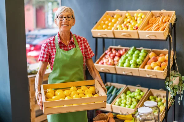 Mature woman works in fruits and vegetables shop. She is holding basket with lemons.